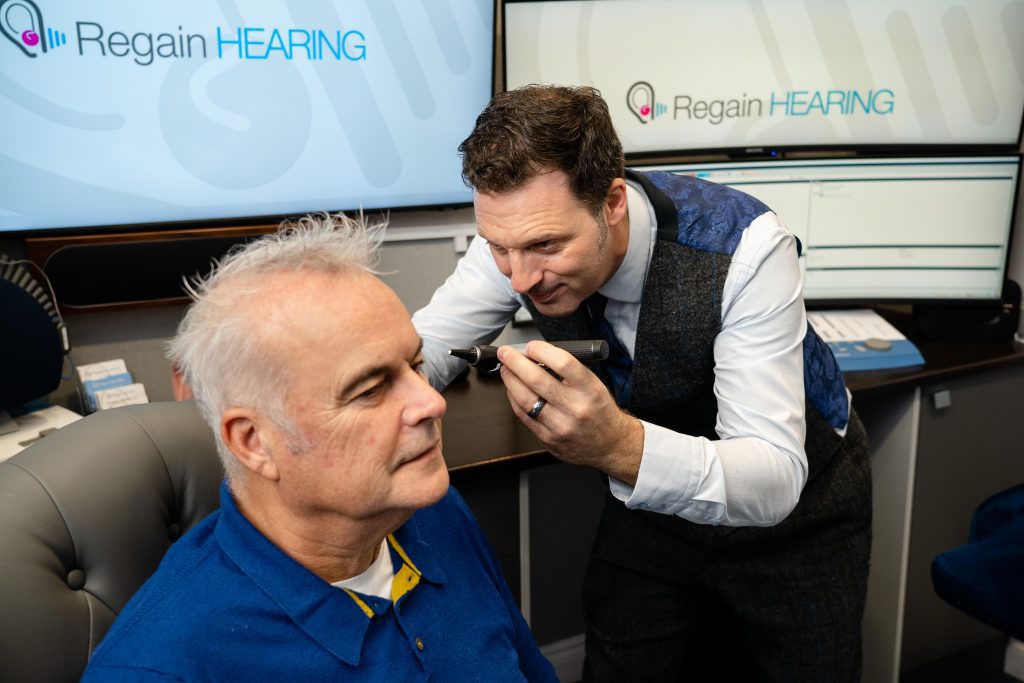 A hearing specialist examines an older mans ear with a medical device in a clinic. The man sits in a chair, and “Regain HEARING” is displayed on screens in the background.