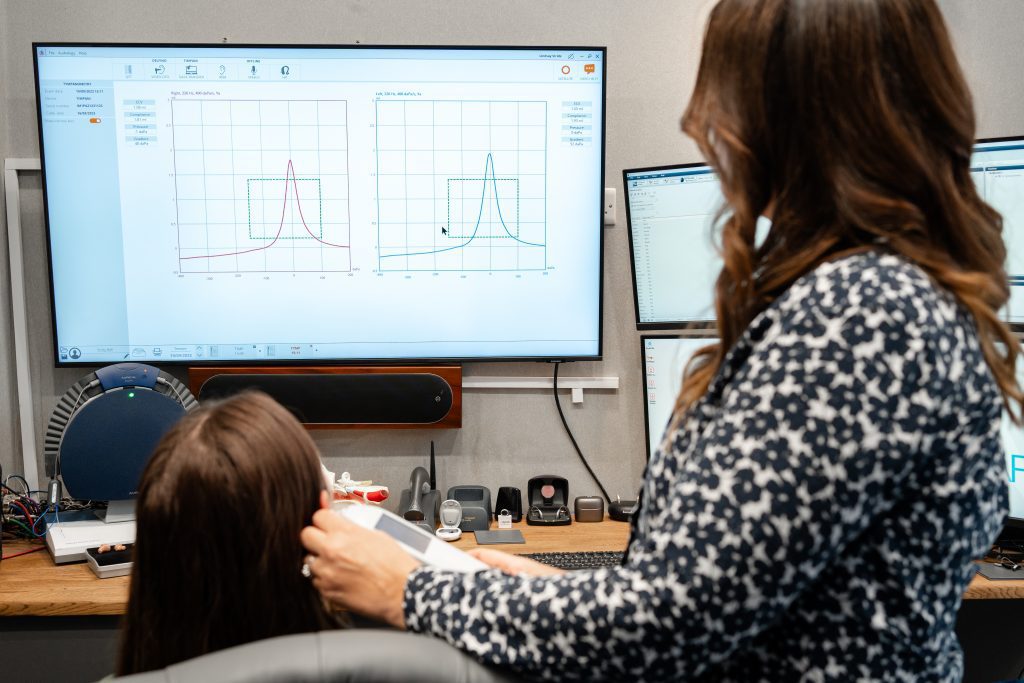 Two women in an office look at graphs displayed on a large computer monitor, with one woman standing and pointing at the screen and the other sitting in front of multiple monitors and technical equipment.