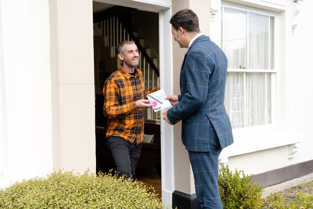 A man in a plaid orange shirt stands at his front door, smiling as he receives papers from a man in a blue checkered suit, who is also smiling. They appear to be engaged in a friendly exchange.