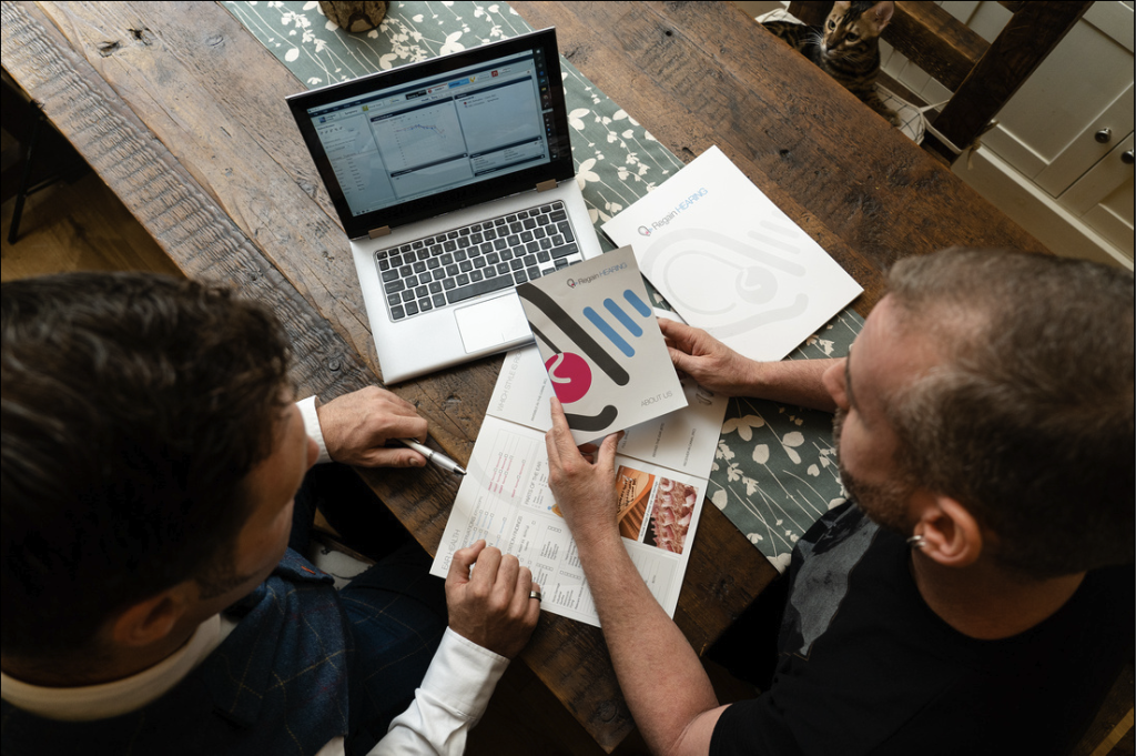 Two people sit at a wooden table reviewing printed documents and a brochure, with an open laptop displaying charts in front of them. One person points at a page, suggesting a business discussion or meeting.