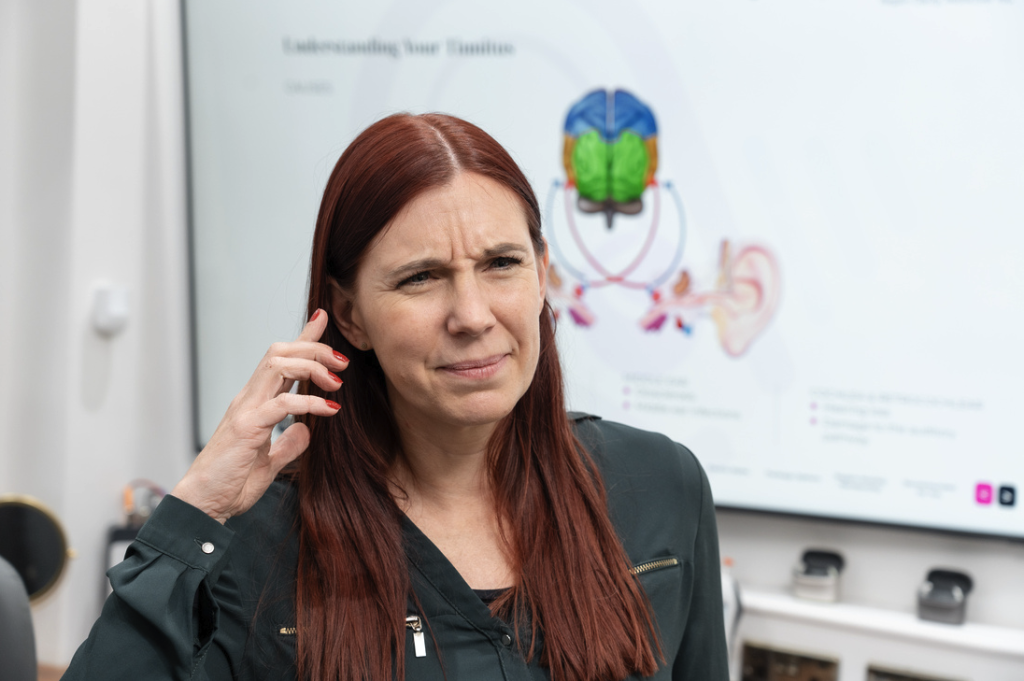 A woman with long red hair touches her ear and looks concerned. Behind her is a screen displaying a diagram of the brain and ear, with the heading “Understanding Your Tinnitus.”.