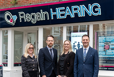 Four professionally dressed people stand smiling outside a storefront with a sign that reads Regain HEARING. The storefront has large windows and informational posters visible inside.