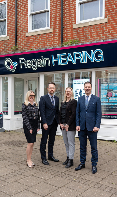 Four people in business attire stand smiling outside a shop with a sign that reads Regain HEARING. The storefront has large windows and a brick exterior.
