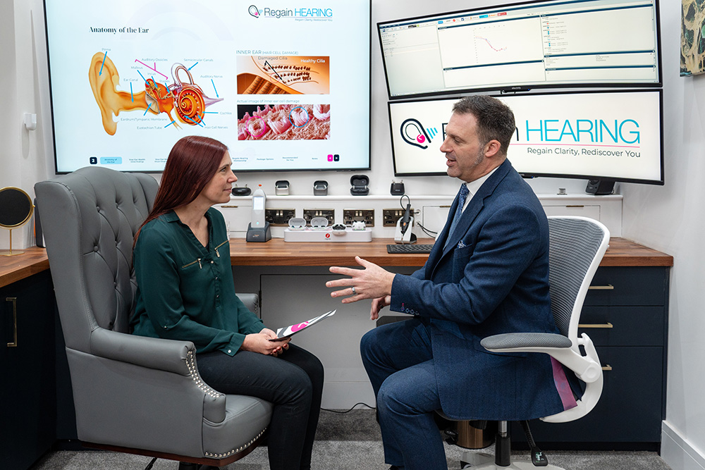 A woman and a man sit facing each other in a modern office, discussing something. Behind them, screens display diagrams of an ear and information about hearing. The office has a professional and welcoming atmosphere.