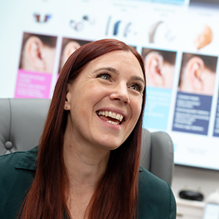 A woman with long red hair smiles while sitting in an office with informational posters and images of ears in the background.