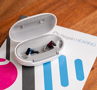 A pair of in-ear hearing aids, one blue and one red, sits in an open white charging case placed on top of a brochure with the words Regain HEARING visible on a wooden table.