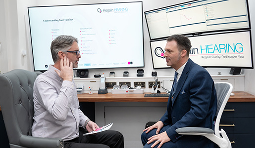 Two men sit facing each other in a modern office. One man gestures toward his ear while the other listens attentively. Behind them are screens and charts related to hearing, with the words Regain HEARING displayed.